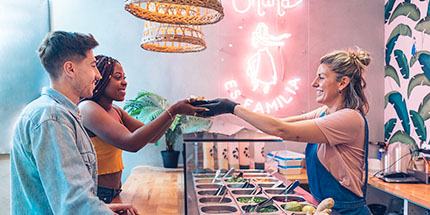 franchise A woman working a buffet style line in a restaurant cheerfully hands food over a sneeze guard to a female customer while a male customer looks on